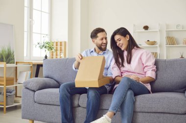 Genç mutlu çift evdeki oturma odasındaki kanepede oturan karton kutuyu açıyor. Erkek ve kadın alıcılar paketleri açıyorlar ve online sipariş alıyorlar. Teslimat ve alışveriş online konsepti.