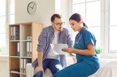 Tıp doktorlarının ofisindeki genç adam klinikte muayene sırasında randevu ve danışmanlık ile rapor tutan dost canlısı bir kadın hemşireyi dinliyor. Tıp konsepti.