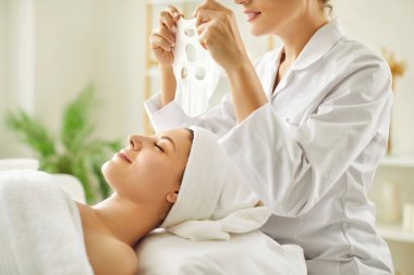 Güzellik uzmanı ya da kozmetikçi, güzellik salonundaki kırışıklık önleyici kozmetik işlemleri için genç kadın yüzüne nemlendirici çarşaf maskesi uyguluyor. Kozmetoloji ve profesyonel cilt bakımı kavramı.