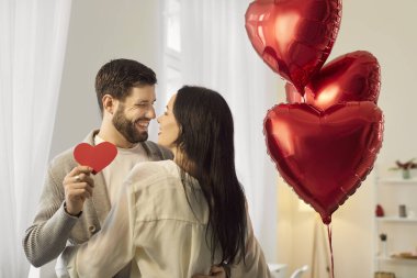 Olumlu evli çiftler sevgililer gününü evde kutluyor, ellerinde kırmızı bir kalple birbirlerine sarılıyorlar. Bu tatil boyunca aşk ve romantizm parlıyor, aile için özel bir an..