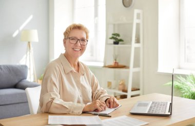 Ev ofisinden bilgisayarla çalışan ve kameraya bakan yaşlı bir kadının portresi. Mutlu kadın muhasebeci, üretken ve neşeli bir çevreyi kucaklayarak görevlerine odaklanıyor..