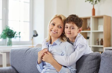 Oturma odasında küçük çocuğuyla koltukta oturan ve neşeli bir şekilde kameraya bakan mutlu bir annenin portresi. Aile, sevgi, ilgi ve anneler günü konsepti.
