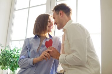 Genç, neşeli bir kadın ve kırmızı kalbi elinde tutan bir adam. Pencere kenarında durup birbirlerine bakıyorlar. Aşık mutlu bir çift. Romantik, ilişki ve Sevgililer Günü konsepti.