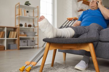 Kırık bacaklı yaşlı bir adam telefonda konuşurken evdeki bir kanepede dinleniyor. Travma tedavisi, koltuk değneklerini gösteren ve yaralanmaya rağmen günlük aktiviteleri yöneten adam..