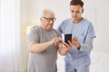 Pozitif doktor ya da bakıcı yaşlı bir adama, huzurevindeki bir hastaya telefonu anlaması ve kullanması için yardım ediyor. Bakım ortamında çevrimiçi teknoloji ve internet navigasyonu.