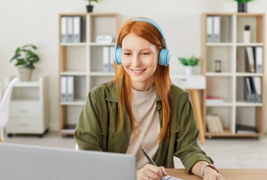 Genç, kızıl saçlı bir kız öğrencinin portresi iş yerinde günlük giysiler ve kulaklıklar takıyor dizüstü bilgisayarla çalışıyor ve not defterine not alıyor. Çevrimiçi eğitim veya uzaktan çalışma kavramı