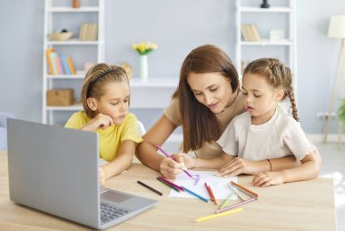 Genç ve mutlu bir annenin portresi. Bilgisayarlı ve çocuklu bir masada oturuyor ve online derste evde resim çiziyor. Aile eğlencesi ve e-eğitim kavramı.
