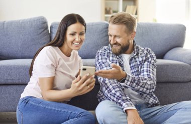 Neşeli Kafkas aile çifti kanepenin yanında oturuyor, cep telefonlarını evde eğlenmek için birlikte kullanıyorlar. Mutlu karı koca internette sörf yaparken ya da komik videolar izlerken gülüyor.