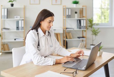 Genç, gülümseyen bir kadın doktorun portresi. Çalışma masasında oturuyor ve klinikte çalışıyor. Ofiste steteskop takan kadın doktor dizüstü bilgisayar ve daktilo kullanıyor..