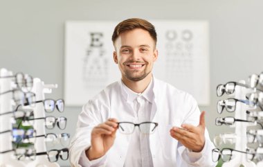 Gülümseyen genç göz doktoru optik mağazada ya da oftalmoloji kliniğinde yeni gözlük gösteriyor. Görme yetisini geliştirmek için gözlük sunan profesyonel gözlükçü, gözlük seçiminde yardımcı olmaya hazır.