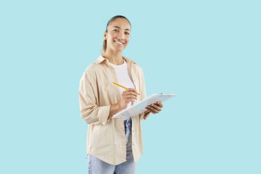 Mutlu, çekici bir kadın panoya yazıyor, gönüllü teşvik ediyor, dostça gülüşünü sorguluyor, pastel ışıkta izole edilmiş soluk turkuaz arka plan, stüdyo fotoğrafı için kameraya poz veren bir model.