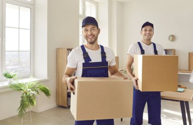 Taşıma servisi. Taşımacılık servisinden iki neşeli erkek hamal karton kutular taşıyorlar. Tulum giymiş gülümseyen profesyonel nakliyecilerin portresi müşterinin daireden ya da ofisten taşınmasına yardımcı oluyor.