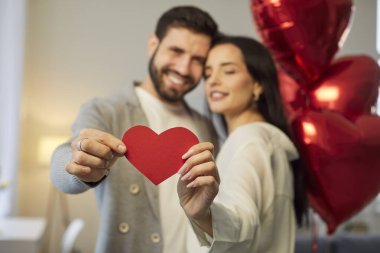 Genç esmer adam ve kadın ellerinde kalp tutuyorlar ve arka planda tatil kutlayan kırmızı folyo balonlarla sarılıyorlar. Aşık mutlu bir çift. Dikkat, şefkat ve sevgililer günü konsepti.