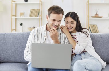 Mutlu gülümseyen genç çift dizüstü bilgisayar planlama bütçesini izliyor, e-postada iyi haberler okuyor, kadın ve erkek bilgisayar ekranına bakıyor, mali durumlarını kontrol ediyor, evdeki kanepede birlikte oturuyor, rahatlıyor. 