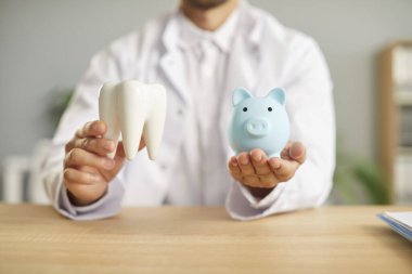 Diş hekimliği kliniğindeki masada oturan, kumbarayı elinde tutan ve sağlıklı beyaz dişleri olan bir dişçinin portresi. Pahalı diş tedavisi için para biriktirme kavramı