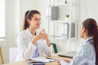 Beyaz üniformalı genç bayan doktor hastane masasında oturmuş konuşuyor, danışmanlık yapıyor, modern tıp kliniğinde danışmanlık yapıyor, sağlık hizmetlerini destekliyor, randevu alıyor.