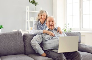 Yaşlı gri saçlı yaşlı çift evdeki oturma odasında kanepede otururken dizüstü bilgisayar kullanıyor ve gülümsüyor. Olgun erkek ve kadın kameraya neşeli görünüyor. E-öğrenme ya da uzaklık serbest çalışma