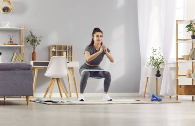 Kadın, ev çalışmasının bir parçası olarak esneme bandıyla çömelme hareketleri yapıyor. Ev ortamında spor ve spor eğitimi rutinini korumak için güç egzersizlerinde aktif kalıyor..