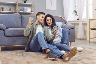 Akıllı telefonlu mutlu genç bir çift, karı koca oturma odasında dinleniyor. Mutluluktan havalara uçuyorlar. İnternetten piyango kazanıyorlar.