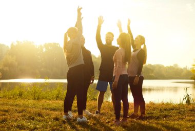 Spor kıyafetli bir grup insan göl kıyısında bir çember oluşturarak parktaki gün batımında doğa çalışmasından sonra ellerini kaldırdılar. Açık hava eğitimi, spor konsepti..