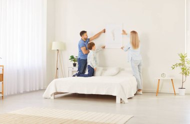 Mutlu bir aile, oğlu rahat bir yatak odasının duvarına çerçeveli bir resim asıyor ve eve kişisel bir dokunuş ekliyor. Mutlu takım çalışması, yenileme ve sıcak bir ortamda paylaşılan dekorasyon..