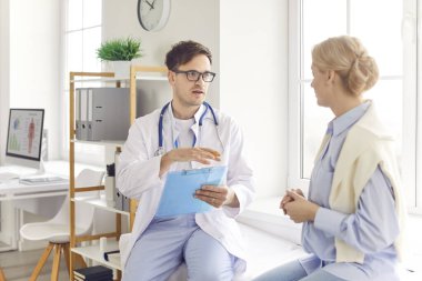 Tıp bürosunda kanepede oturan genç kadın hasta ellerinde randevu ile rapor tutan ve klinikteki muayene sırasında danışmanlık yapan dost canlısı erkek doktor terapistleri dinliyor..