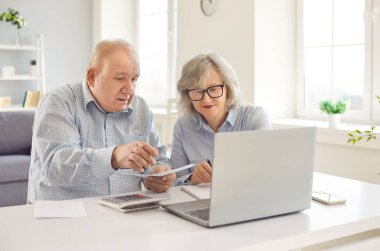 Online faturaları ödeyen yaşlı çift. Eski karı koca hesap makinesi ve modern dizüstü bilgisayarla masada oturuyorlar, aile bütçesini yönetiyorlar, faturaları ödüyorlar, masrafları planlıyorlar.