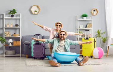 Komik, mutlu şapkalı ve güneş gözlüklü çift evde bavullarıyla tatil için hazır şişme yatakta otururken eğleniyor. Tatil ve yaz yolculuğu konsepti.
