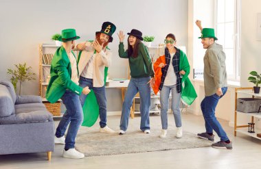 Happy smiling young joyful people friends men and women in funny green clothes and hats dancing at party having fun in the living room at home celebrating Saint Patricks Day together.