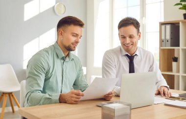 İki genç kendine güvenen iş adamı ve şirket çalışanı ofiste dizüstü bilgisayarla çalışıyor ve iş yerindeki mali belgelere bakıyor iş projelerini tartışıyorlar..