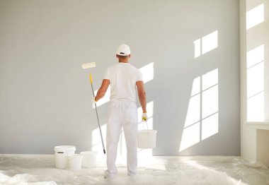 Beyaz üniformalı bir erkek ressamın elinde kova ve elinde boya silindiri boş bir odada gri bir duvarı boyamaya giderken görüntüsü. Yenileme, apartman onarımı ve yenileme konsepti..