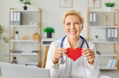 Kardiyolojide uzman yaşlı bir kadın kardiyolog ya da hemşire portresi, elinde kırmızı bir kalp tutuyor. Hastane ortamında sağlık, şefkat ve mesleki adanmışlık.