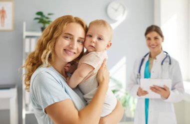 Çocuk doktorunun muayenehanesinde mutlu sağlıklı anne ve bebek. Çocuk kliniğinde küçük çocuğu olan genç bir kadın. Portre güzel gülümseyen anne kucağında çocuk kameraya bakıyor, doktor arka planda.