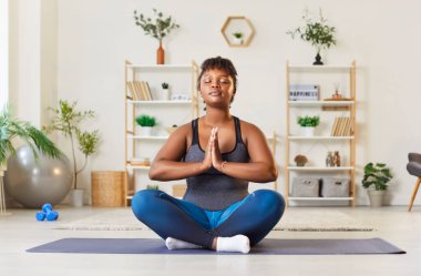 Barışçıl Afro-Amerikalı kadın evde spor yaparken zihnini ve vücudunu ferahlatmak için lotus pozisyonunda meditasyon yapıyor. Yoga pratiği rahatlama ve aktif eğitim dengesini yansıtıyor..