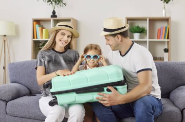 Şapkalı mutlu ebeveynler ve güneş gözlüklü kızları kanepede dizlerinin üstünde bavullarıyla birlikte oturuyorlar. Neşeli aile valizlerini topluyor, yaz yolculuğuna hazırlanıyor. Aile tatili kavramı.