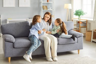 Neşeli, sevgi dolu anne, gri kanepede, aydınlık oturma odasında oturuyor, eğleniyor, gülüyor ve iki neşeli ve büyüleyici çocuğuyla kaliteli vakit geçiriyor, birlikte mutlu aile anıları yaratıyor..