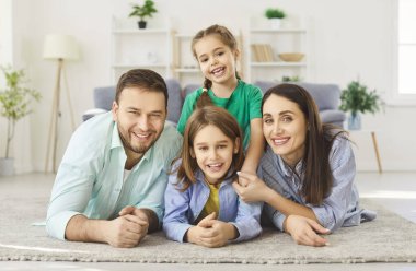 Gülümseyen dört çocuklu mutlu aile, iki çocuk evde yerde yatıyorlar ve kameraya neşeli bir şekilde bakıyorlar. Anne ve babanın çocuklarla vakit geçirdiği portre. Mutlu aile kavramı.