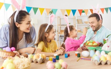 Mutlu gülümseyen aile Paskalya hazırlıkları sırasında yumurta boyarken eğleniyor. Anne, baba ve tavşan kulağı takan iki komik çocuk mutfakta oturmuş Paskalya bayramını kutluyorlar..
