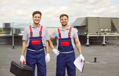 Çatıda bir havalandırma sisteminin bakımını ve tamirini yapan gülümseyen iki elektrikçinin portresi. Profesyoneller hizmet ve kurulum, uzmanlık ve takım çalışması sergilemekle meşgul.