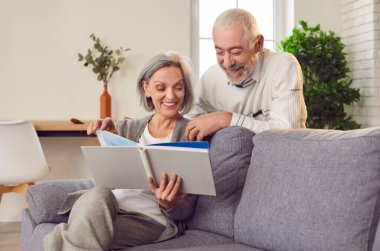 Kanepede oturan mutlu yaşlı çift aile albümüne bakıyor. Yaşlı çift aile fotoğrafı albümüne bakıyor. Yüzlerinde mutlu bir gülümseme beliriyor. Yaşlı çift aile fotoğrafı albümüne bakıyor.