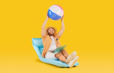 Memnun adam dizüstü bilgisayar ve plaj topu ile rahatlıyor, plajda yaz tatilinin tadını çıkarıyor. Onun mutlu ifadesi turuncu bir arka planda geçen seyahat ve gevşemeyi yansıtıyor..
