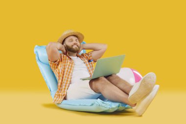 Rahat giyinen, şişme yatakta oturan, gözleri kapalı çalışan ya da sarı arka planda internetten bilet alan komik bir adam. Yaz tatili, tatil gezisi ya da uzaktan çalışma konsepti