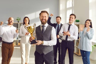 Mutlu gülümseyen genç bir adamın portresi. İş hayatında başarı ödülünü elinde tutuyor. Meslektaşlarından oluşan bir ekip şirketin en iyi çalışanı ödülünü alkışlıyor..