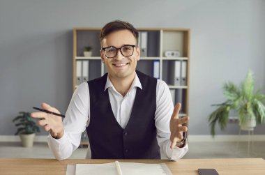 Ofiste çalışan genç ve çekici bir iş adamı kameraya bakıyor ve iş yerindeki masada video görüşmesi yapıyor. Online sohbet eden ya da görüntülü görüşme yapan kendinden emin bir adam..