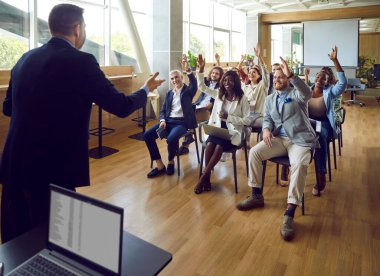 Modern ofisteki iş toplantısında soru sormak için çok ırklı genç meslektaşlar el kaldırıyor. Toplantı odasında çeşitli iş adamları konuşmacılarını dinlerken konferansta oy veriyorlar..