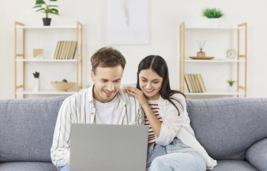 Dizüstü bilgisayarda genç bir çift, bilgisayar ekranında izleme, bütçe planlama, son dakika haber maillerini okuma, gülümseyen kadın ve erkek mali durumlarını kontrol etme, evdeki kanepede birlikte oturma, internet başvurusu.