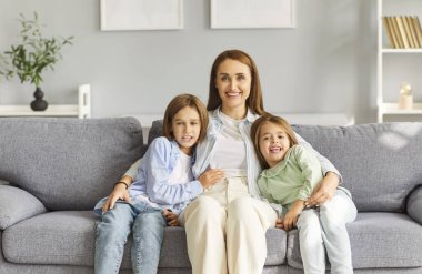 Mutlu gülümseyen bir aile, iki küçük sevimli çocuğu olan bir anne kameraya bakıyor, gülüyor, eğleniyor, evdeki yumuşak kanepede birlikte ilgileniyorlar, rahat hissediyorlar, evde güvendeler.