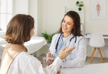 Steteskoplu, beyaz üniformalı gülümseyen doktor portresi randevu ile rapor tutuyor ve klinikteki tıbbi muayene sırasında bir kadın hastaya danışmanlık yapıyor..