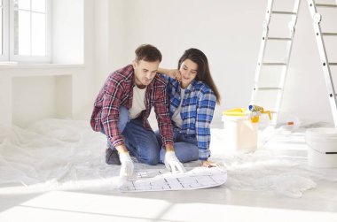 Parlak odada yerde oturan mutlu çift inşaat planlarına ve yenileme planlarına bakıyor. Gelişigüzel giyinen genç bir aile ev geliştirme projesi sırasında iç tasarım fikirlerini tartışıyor..