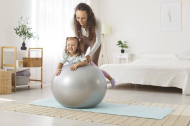 Gülümseyen genç anne, parlak ferah yatak odasında spor salonundaki mutlu küçük kızını kucaklıyor. Neşeli bir aile babası ve çocuk evde spor yaptıktan sonra eğleniyorlar. Aile fitness konsepti.